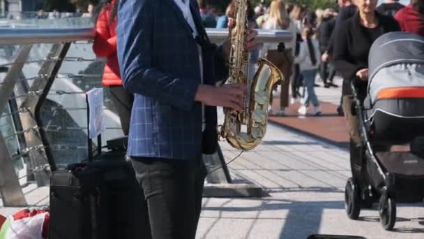 Młody muzyk uliczny gra na saksofonie w parku publicznym, ludzie chodzą przeszłością — Wideo stockowe