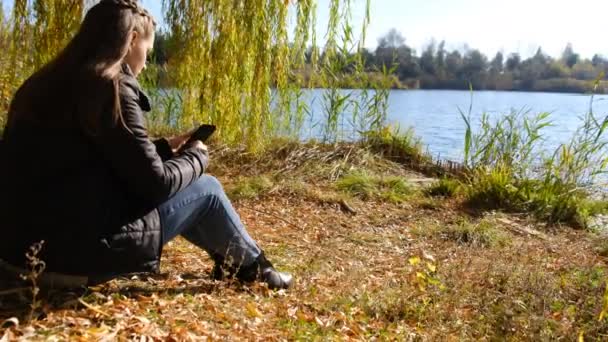 Junge Frau mit Smartphone in der Hand sitzt im Herbst auf gelbem Laub am Fluss — Stockvideo