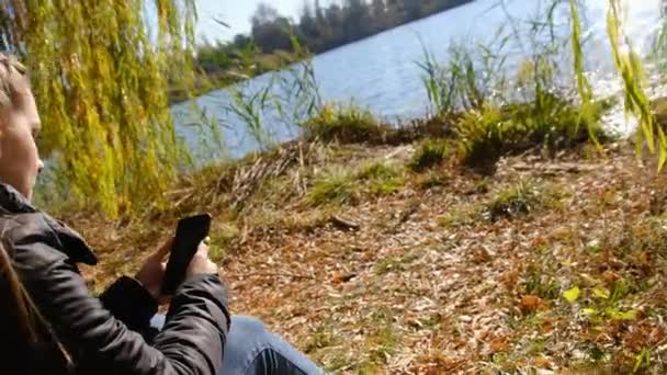 Junge Frau mit Smartphone in der Hand sitzt im Herbst auf gelbem Laub am Fluss — Stockvideo