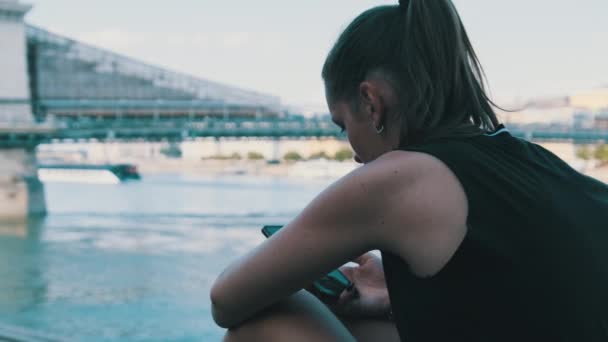 Молода жінка користується смартфоном на узбережжі біля річки — стокове відео