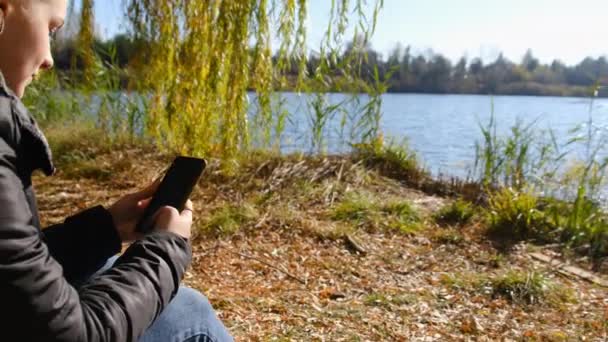 Junge Frau mit Smartphone in der Hand sitzt im Herbst auf gelbem Laub am Fluss — Stockvideo