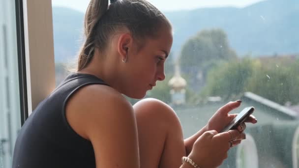 Mulher bonita com Smartphone Senta-se no peitoril da janela pela janela na cidade — Vídeo de Stock