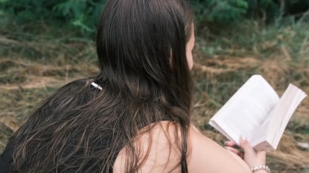 Mujer joven lee un libro sentado en la hierba en la naturaleza en el día de verano — Vídeos de Stock