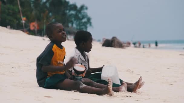 Dos chicos africanos locales se sientan en la playa y tocan tambores improvisados de botella, Zanzíbar — Vídeo de stock