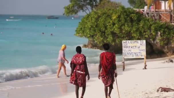Masai, Tanzanya 'nın Zanzibar kentindeki turistler arasında okyanus kıyısındaki sahil boyunca yürüyor. — Stok video