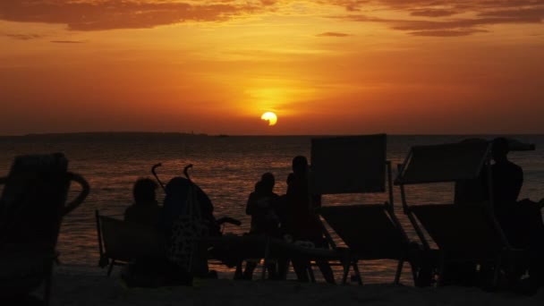 Silhuetas de pessoas ao pôr do sol tendo um descanso à beira-mar na praia, Zanzibar — Vídeo de Stock