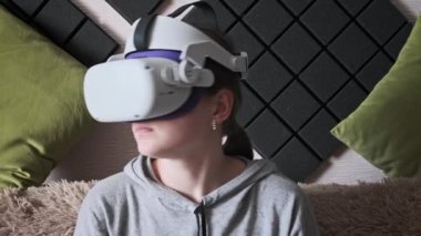Genç bir kadın içeride video kask kullanarak oyun oynuyor. Sanal gerçeklik gözlüklü kız kanepede oturuyor ve evdeki kontrolörleri hareket ettiriyor. VR simülatörü takıyor, 3 boyutlu siber uzayın tadını çıkarıyor. Artırılmış gerçeklik