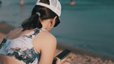 Genç bir kadın sahilde akıllı telefon kullanıyor. Plajda oturan mayo giymiş güzel bir kız elinde bir cep telefonu tutuyor ve sosyal ağları izliyor. Yaşam tarzı. Deniz kıyısındaki eğlence aktiviteleri