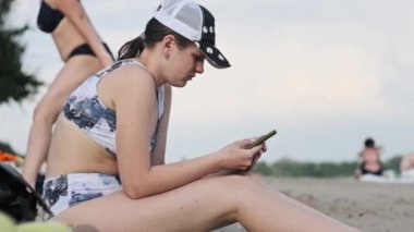 Mayolu güzel bir kız kumlu bir sahilde elinde telefonla oturuyor. Genç kadın akıllı telefon kullanıyor. Güzel bir kadın cep telefonu tutuyor ve sosyal ağları izliyor. Yaşam tarzı. Boş zaman aktiviteleri