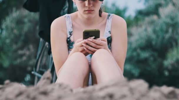 若い女性はビーチでスマートフォンを使用しています ビーチに座っている水着の美しい女の子は 彼女の手の中に携帯電話を保持し ソーシャルネットワークを見ています ライフスタイル 海岸でのレジャー活動 — ストック動画
