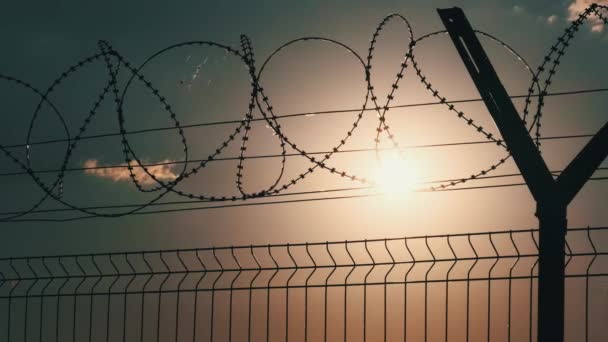 日没に対するフェンスの上に有刺鉄線 夕日はフェンスの上にかみそり線の後ろに輝いています オレンジの空に国境フェンスのシルエット 上部に有刺鉄線付きチェーンリンクフェンス — ストック動画