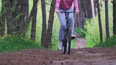 Young woman on a bicycle rides along a forest path in summer day, slow motion. Woman on bike riding on a trail way between green trees in a countryside park. Sport lifestyle, leisure activity, holiday