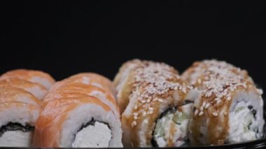 Suşi plastik kutuda yuvarlanır, siyah arka planda döner, yakın plan. Philadelphia peynirli taze Japon suşisi, nori, susam, somon filetosu pilav, avokado ve salatalık. Plastik kabın içindeki suşi.