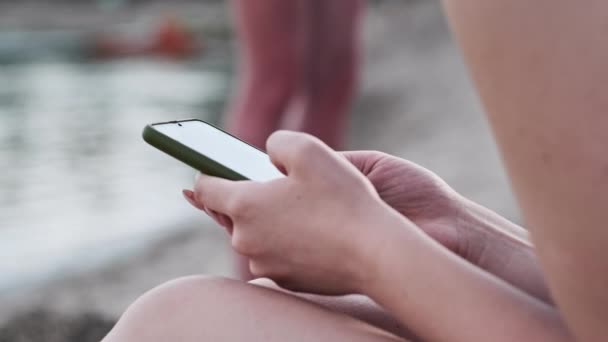 水着姿の美少女が携帯電話を手に砂浜に腰を下ろしている 若い女性はスマートフォンを使う きれいな女性は携帯電話を保持し ソーシャルネットワークを監視します ライフスタイル レジャー活動 — ストック動画