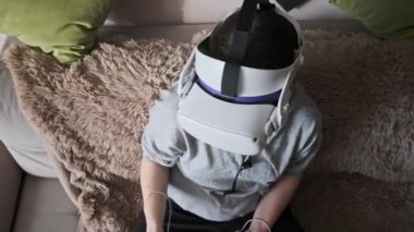 Sanal gerçeklik kaskı takan genç bir kadın kanepede oturuyor ve evde elinde joysticklerle oturuyor. Kapalı alanda video kask kullanarak oyun oynamak. VR simülatörü takan kız 3 boyutlu siber uzayın tadını çıkarıyor. VR gözlükleri