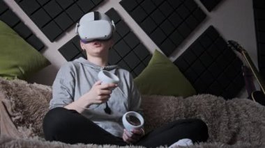 Sanal gerçeklik kaskı takan genç bir kadın kanepede oturuyor ve evde elinde joysticklerle oturuyor. Kapalı alanda video kask kullanarak oyun oynamak. VR simülatörü takan kız 3 boyutlu siber uzayın tadını çıkarıyor. VR gözlükleri