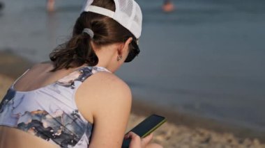 Mayolu güzel bir kız kumlu bir sahilde elinde telefonla oturuyor. Genç kadın akıllı telefon kullanıyor. Güzel bir kadın cep telefonu tutuyor ve sosyal ağları izliyor. Yaşam tarzı. Boş zaman aktiviteleri