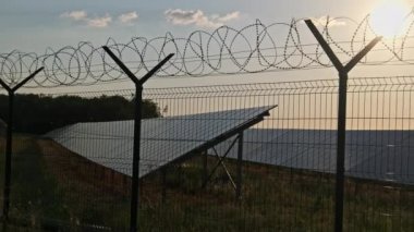 Güneş enerjisi santralinin etrafında gün batımında dikenli teller var. Çit örgüsü. Güneş enerjisi elementlerinin bulunduğu bölgeyi çitliyor. Dikenli tellerin arkasındaki güneş panelleri alanı.