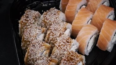 Plastik kutuda suşi yuvarlanıyor, yakın plan. Philadelphia peyniri, nori, susam, somon filetosu, pirinç, avokado ve salatalıklı taze Japon suşisi. Plastik kabın içindeki suşi..