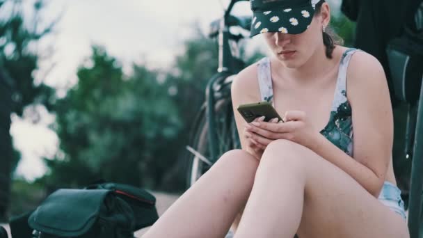 若い女性はビーチでスマートフォンを使用しています ビーチに座っている水着の美しい女の子は 彼女の手の中に携帯電話を保持し ソーシャルネットワークを見ています ライフスタイル 海岸でのレジャー活動 — ストック動画