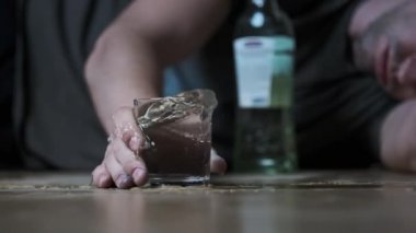 Sarhoş bir adam bir bardak votka ve aşırı dozda alkolle yere düşüyor. Ağır çekim. Alkol bağımlılığı. Sarhoş bir gencin eli şişenin yanına düştü. Yakın plan. Alkolizm, Bağımlılık