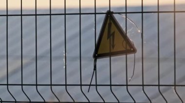 Güneş santralinin tel örgüsünde yüksek voltaj işareti var. Sarı üçgen şimşek işareti yüksek voltaj tehlikesi. Elektrik santralinin elektrik güvenlik sembolü uyarısı. Elektrik şoku tehlikesinin işareti.
