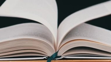 Kitabın sayfaları yavaş çekimde kendi kendine dönüyor, yakın çekimde. Beyaz kitabın sayfaları rüzgarın etkisiyle rastgele yer değiştiriyor. Stüdyoda siyah arka plan üzerine açık bir kitap