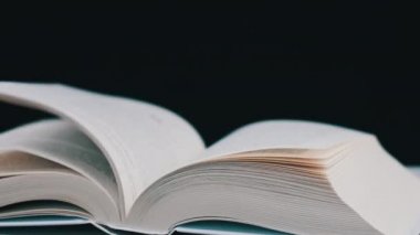 Kitabın sayfaları yavaş çekimde kendi kendine dönüyor, yakın çekimde. Beyaz kitabın sayfaları rüzgarın etkisiyle rastgele yer değiştiriyor. Stüdyoda siyah arka plan üzerine açık bir kitap