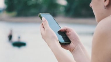 Genç bir kadın sahilde akıllı telefon kullanıyor. Plajda oturan mayo giymiş güzel bir kız elinde bir cep telefonu tutuyor ve sosyal ağları izliyor. Yaşam tarzı. Deniz kıyısındaki eğlence aktiviteleri