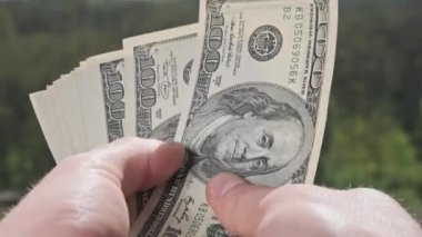 Erkek eller yeşil doğanın arka planına karşı bir yelpaze gibi eski dolarları gösteriyor. Genç adam elinde bir sürü 100 dolarlık banknot tutuyor. Açık hava banknotları yığını. Kâr kavramı, kağıt para biriktirme