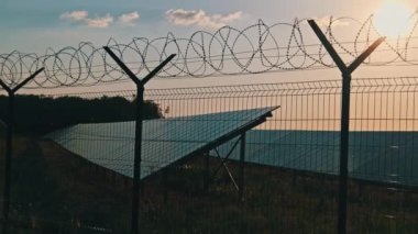 Gün batımında güneş enerjisi santralinin etrafında dikenli teller var. Çit örgüsü. Güneş enerjisi elementlerinin bulunduğu bölgeyi çitliyor. Dikenli tellerin arkasındaki güneş panelleri alanı.
