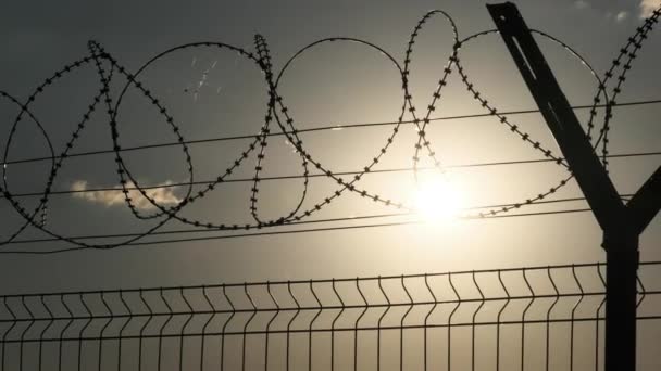 夕阳西下栅栏上有铁丝网 夕阳在铁丝网后面的篱笆上闪烁着光芒 橙色天空下的边境围栏轮廓 顶部有带刺铁丝网的链条围栏 — 图库视频影像