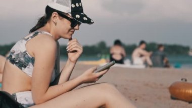 Genç bir kadın sahilde akıllı telefon kullanıyor. Plajda oturan mayo giymiş güzel bir kız elinde bir cep telefonu tutuyor ve sosyal ağları izliyor. Yaşam tarzı. Deniz kıyısındaki eğlence aktiviteleri