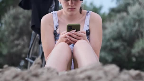 水着姿の美少女が携帯電話を手に砂浜に腰を下ろしている 若い女性はスマートフォンを使う きれいな女性は携帯電話を保持し ソーシャルネットワークを監視します ライフスタイル レジャー活動 — ストック動画