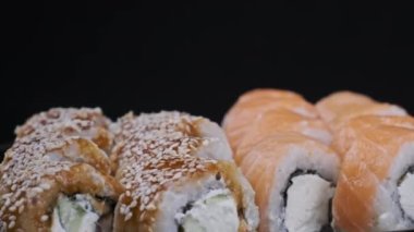 Suşi plastik kutuda yuvarlanır, siyah arka planda döner, yakın plan. Philadelphia peynirli taze Japon suşisi, nori, susam, somon filetosu pilav, avokado ve salatalık. Plastik kabın içindeki suşi.