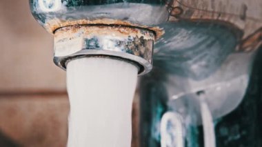 Ağır çekimde akan suyla musluk, makro çekim. Kirlenmiş eski bir musluktan kalsiyum ve kirle dolu bir su fışkırarak banyodaki bir lavaboya akıyor. Su açılıp kapanıyor. Yakın plan..