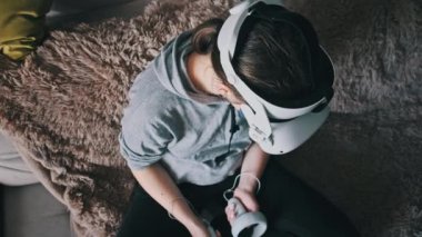 Sanal gerçeklik kaskı takan genç bir kadın kanepede oturuyor ve evde elinde joysticklerle oturuyor. Kapalı alanda video kask kullanarak oyun oynamak. VR simülatörü takan kız 3 boyutlu siber uzayın tadını çıkarıyor. VR gözlükleri