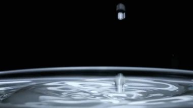 Su damlası su yüzeyine ağır çekimde siyah arka planda düşer, makro çekim. Su yüzeyine çarptıktan sonra farklı daireler oluşur. Sıvının yüzey geriliminin etkisi. 240 fps.