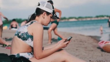 Mayolu güzel bir kız kumlu bir sahilde elinde telefonla oturuyor. Genç kadın akıllı telefon kullanıyor. Güzel bir kadın cep telefonu tutuyor ve sosyal ağları izliyor. Yaşam tarzı. Boş zaman aktiviteleri