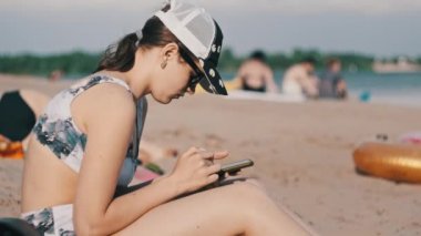 Genç bir kadın sahilde akıllı telefon kullanıyor. Plajda oturan mayo giymiş güzel bir kız elinde bir cep telefonu tutuyor ve sosyal ağları izliyor. Yaşam tarzı. Deniz kıyısındaki eğlence aktiviteleri