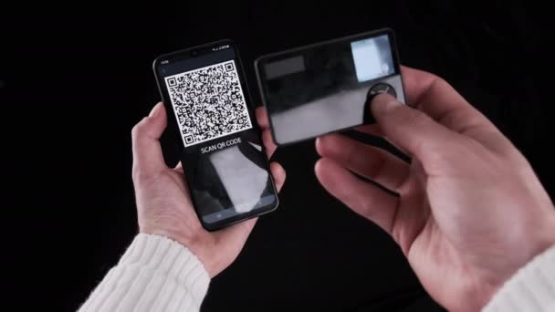 若い男は取引のためにスマートフォン上のQrコードをスキャンするためにポータブルコールド暗号化財布を使用しています 黒の背景にBitcoinを送信するための最新のデバイスカメラを使用しています デジタルキャッシュ 将来の概念 — ストック動画