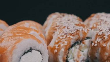 Suşi plastik kutuda yuvarlanır, siyah arka planda döner, yakın plan. Philadelphia peynirli taze Japon suşisi, nori, susam, somon filetosu pilav, avokado ve salatalık. Plastik kabın içindeki suşi.
