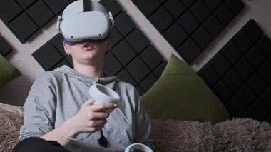 Sanal gerçeklik kaskı takan genç bir kadın kanepede oturuyor ve evde elinde joysticklerle oturuyor. Kapalı alanda video kask kullanarak oyun oynamak. VR simülatörü takan kız 3 boyutlu siber uzayın tadını çıkarıyor. VR gözlükleri