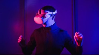 Man in a virtual reality helmet illuminated in red and blue plays a game. Young man in VR headset moves his hands in virtual space. Futuristic technology to play simulation 3D video games Metaverse 4K