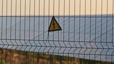 Güneş santralinin tel örgüsünde yüksek voltaj işareti var. Sarı üçgen şimşek işareti yüksek voltaj tehlikesi. Elektrik santralinin elektrik güvenlik sembolü uyarısı. Elektrik şoku tehlikesinin işareti.