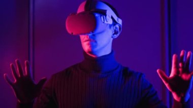 VR kasklı genç adam ellerini kullanarak sanal gerçeklikle etkileşime giriyor. Erkek oyun oyunları, kırmızı-mavi ışık altında VR kulaklıkla 360 medya içeriği. Fütürist teknoloji. Meta-evren kavramına daldırma
