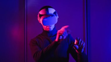 Young man in a VR helmet interacts with virtual reality and media content. Concept of male plays game in VR headset at red-blue light. Use virtual reality futuristic technology. Immersion in metaverse