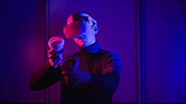 Young man in VR helmet interacts with virtual reality using controllers. Male plays game or see 360 media content in VR headset at red-blue light. Futuristic technology. Concept immersion in metaverse