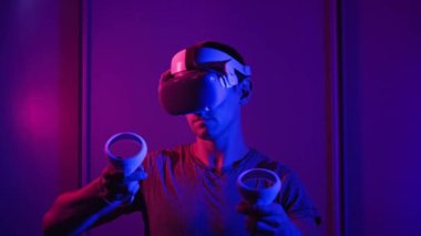 VR kasklı genç adam sanal gerçeklikle kontrolörleri kullanarak etkileşime giriyor. Erkek oyun oynuyor ya da kırmızı-mavi ışıkta VR kulaklıkla 360 medya içeriğini görüyor. Fütürist teknoloji. Meta-evrene daldırma kavramı
