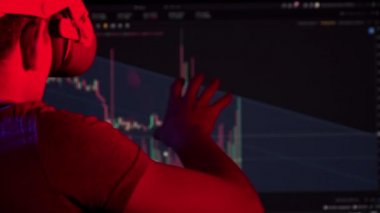 Sanal gerçeklik kaskındaki bir tüccar kripto para çizelgesine bakar. Genç adam kripto para çizelgelerini bir VR kulaklığı ve karanlıkta bir monitör arkaplanı kullanarak analiz ediyor. Geleceğin iş adamı..